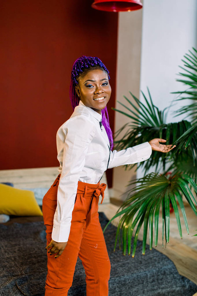 Gorgeous young african american girl , posing indoors, with blue hair. Dressed in white blouse and orange pants. - Photo, Image