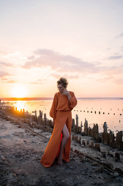 Красивая женщина с элегантными длинными ногами в длинном оранжевом платье на закате. красивые женщины танцуют на закате, восход солнца на берегу моря
 - Фото, изображение