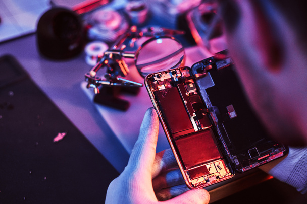 Ο επισκευαστής εξετάζει προσεκτικά την ακεραιότητα των εσωτερικών στοιχείων των το smartphone σε ένα σύγχρονο συνεργείο. Φωτισμός με κόκκινο και μπλε φώτα - Φωτογραφία, εικόνα