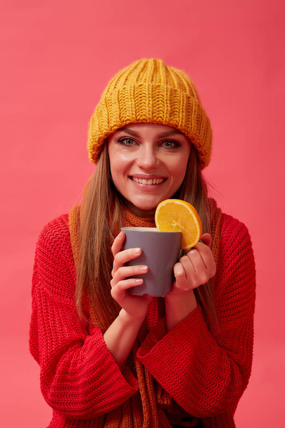 Beautiful girl in warm cozy sweater holds a hot drink with orange. Red background. Studio - Fotografie, Obrázek
