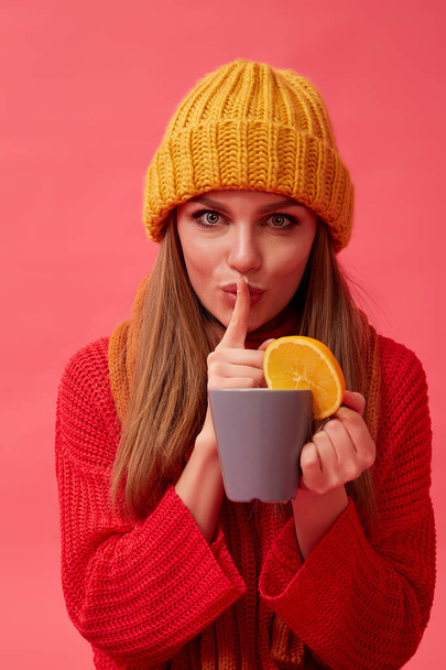 Beautiful girl in warm cozy sweater holds a hot drink with orange. Red background. Studio - Zdjęcie, obraz
