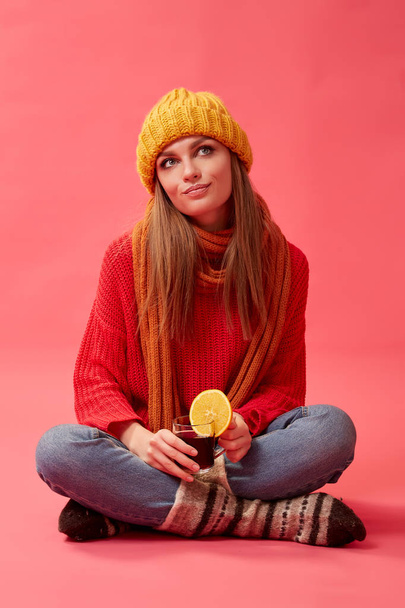 Girl in a red sweater and knitted hat holding a cup of hot mulled wine with an orange slice. Red background. Studio - Fotó, kép