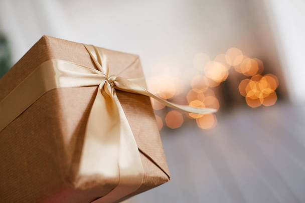 Gift packaging brown tied with a gold ribbon with a bow. Blurred background with lights - Фото, изображение