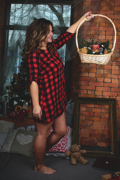 クリスマスツリーの背景にバスケットが付いているシャツの美しい女の子の肖像画. キャンドルとスプルースのスプリッグが付いているバスケット. - 写真・画像