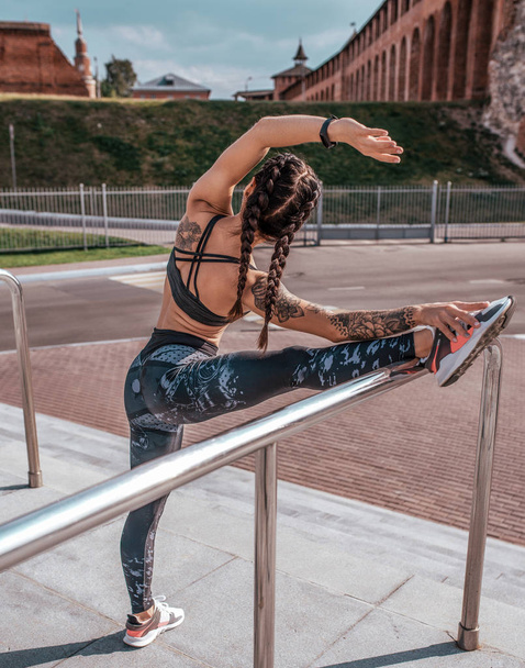 Beautiful young woman in tattoos, summer city, stretches muscles of leg before a workout. in sportswear, leggings and bra. In morning before jogging and fitness in open air. In the splits pose yoga. - Foto, Bild