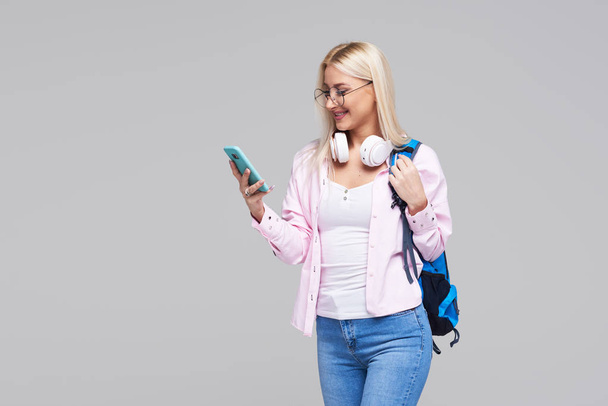 Portret van een student van de vrolijke gelukkig meisje met rugzak, luisteren naar muziek met een koptelefoon en het maken van de selfie op mobiele gadget, telefoon geïsoleerd over grijze achtergrond - Foto, afbeelding