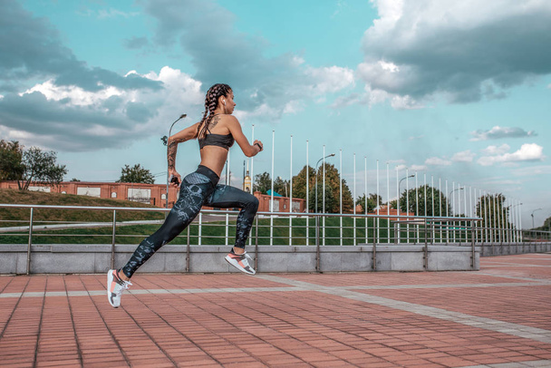 Beautiful young girl tattoos, summer city, runs jump , in hand smartphone listens music in headphones, sportswear leggings bra. Free space text. Morning exercise in fresh air on background blue sky. - 写真・画像