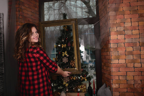 portret van een mooie jonge glimlachende vrouw in een shirt met een fotolijstje op de achtergrond van een raam en een kerstboom en een muur van rode bakstenen - Foto, afbeelding