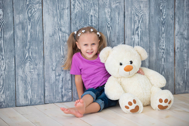 Adorable smiling little girl sitting near a gray wooden wall background and hugging a white teddy bear - Фото, зображення