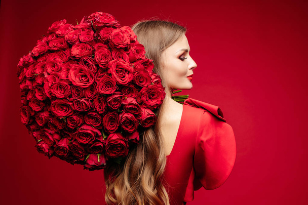 Beautiful young brunette lady in red with bunch of red roses on shoulder. - Photo, Image