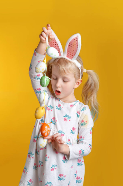 Cute adorable Caucasian blonde girl in white dress with pink  Easter bunny ears playing with eggs in studio on yellow background. Funny kid child celebrating traditional Christian holiday - Photo, Image