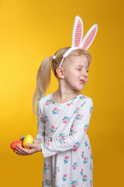 Menina loira branca rabugenta engraçada em vestido branco com orelhas de coelho rosa da Páscoa segurando ovos e se afastando no fundo amarelo. Criança infeliz não está disposta a celebrar feriado cristão
 - Foto, Imagem
