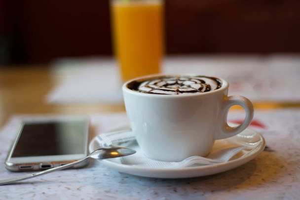 Stillleben mit einer Tasse aromatisiertem Cappuccino, einem Telefon auf dem Hintergrund eines Glases Orangensaft.  - Foto, Bild
