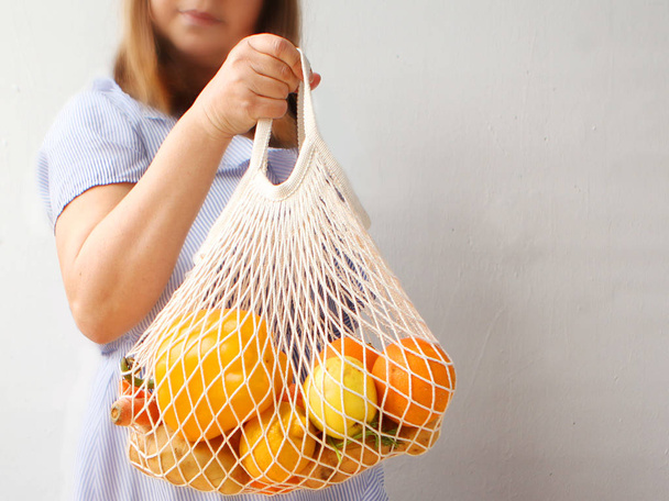 A woman is holding a white mesh bag with fresh vegetables: raw carrots, lettuce, paprika, lemons, greens close-up on a light background - Photo, Image