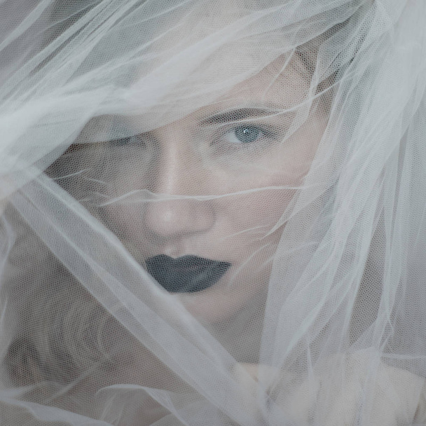 girl's face behind a veil. Portrait of a beautiful blue-eyed girl with black lips behind a white veil. Black lips. tender portrait. Tulle.bewitching glance - Photo, Image