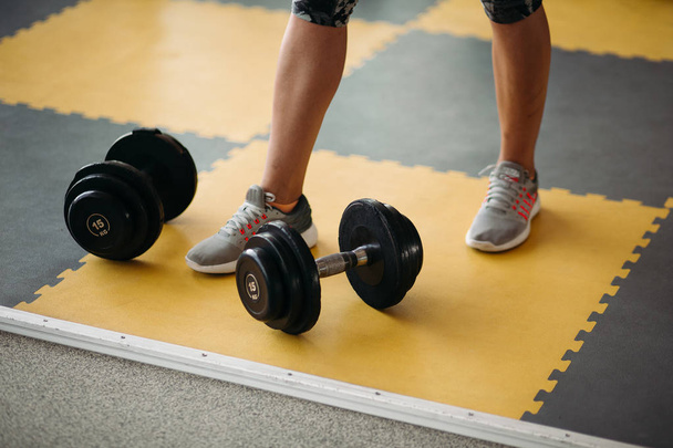 Equipo de fitness dos mancuernas cerca del pie femenino en el suelo amarillo y negro en el gimnasio moderno
. - Foto, Imagen