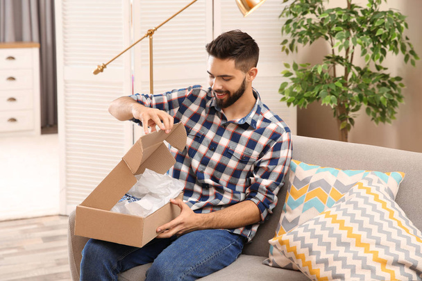 Young man opening parcel on sofa at home - 写真・画像