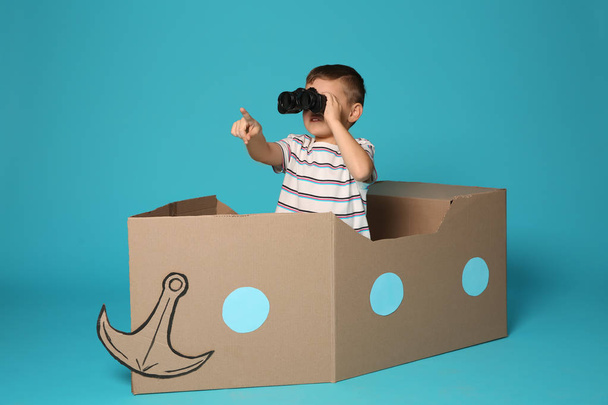 Cute little boy playing with binoculars and cardboard boat on color background - Foto, afbeelding
