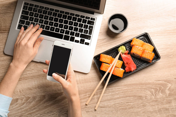 Woman with smartphone, laptop and tasty sushi rolls at wooden table, space for text. Food delivery - Zdjęcie, obraz