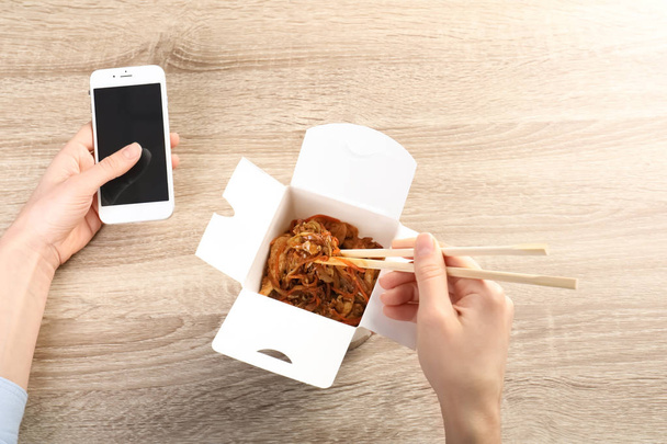 Woman holding smartphone with space for text while eating Chinese noodles at wooden table, closeup. Food delivery - Fotó, kép
