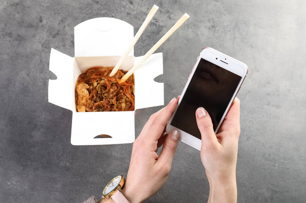 Mulher com smartphone e macarrão chinês na mesa cinza, espaço para texto. Entrega de alimentos
 - Foto, Imagem