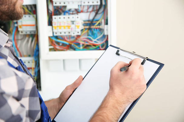 Male electrician with clipboard near fuse board indoors, closeup - Photo, image