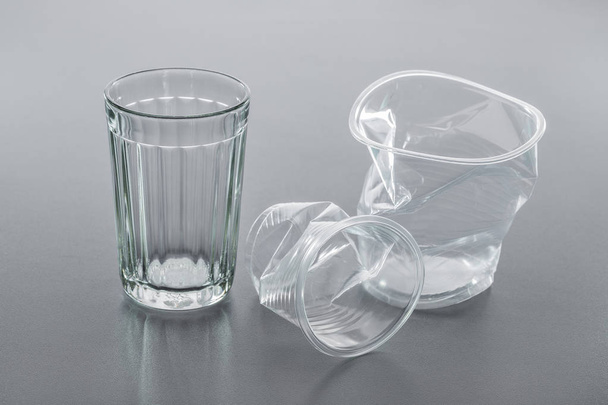 Simple glass and crumpled plastic cup on gray background, close-up. Recycling concept. - Photo, image