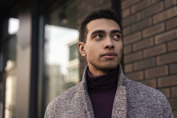 Closeup portrait of handsome African American man with positive face waiting for taxi outdoors. Successful fashion model wearing stylish grey coat and purple turtleneck walking along urban street - Fotografie, Obrázek