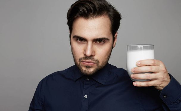 Young confident man holding hand glass of fresh milk on gray background - Foto, Imagem