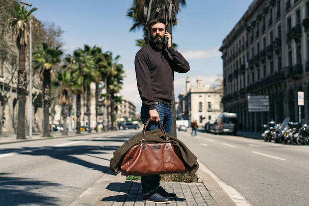 Attractive bearded man holding travel bag and using mobile mobile phone outside. Blurred background. - Foto, Bild
