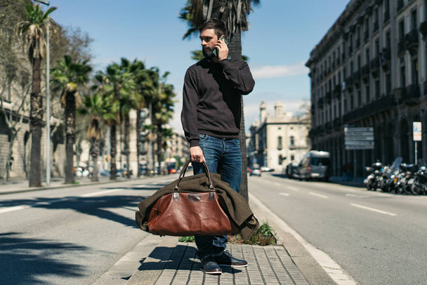Attractive bearded man holding travel bag and using mobile mobile phone outside. Blurred background. - Foto, Imagen