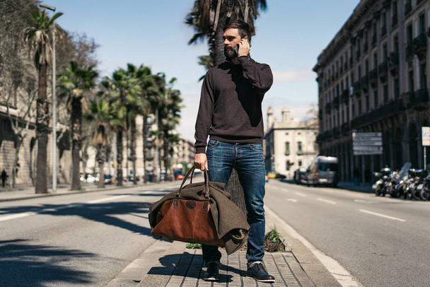 Atractivo hombre barbudo sosteniendo bolsa de viaje y el uso de teléfono móvil fuera. Fondo borroso
. - Foto, Imagen