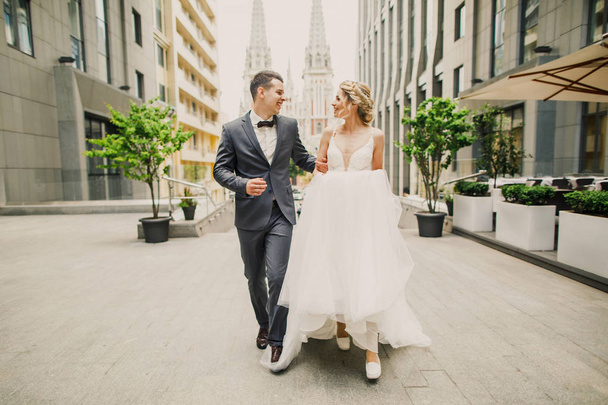 young beautiful couple bride and groom walk in the city on the background of the church and smile the bride in a beautiful dress the groom dressed stylishly - Фото, изображение