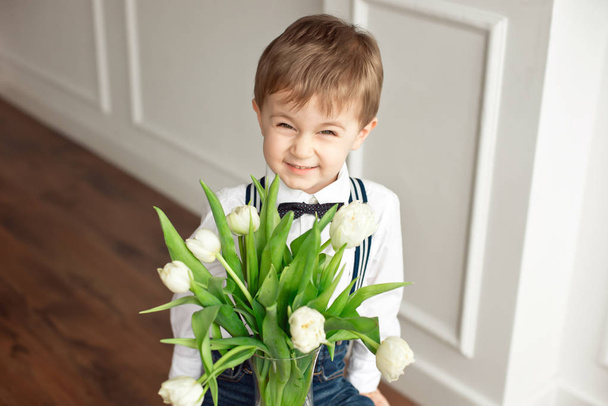 Cute boy in a white shirt and a tassel butterfly gives a bouquet of white tulips. - Foto, Bild