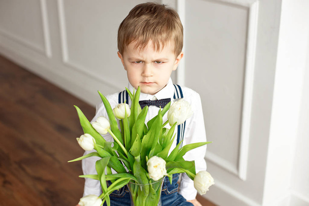 Cute boy in a white shirt and a tassel butterfly gives a bouquet of white tulips. - Zdjęcie, obraz