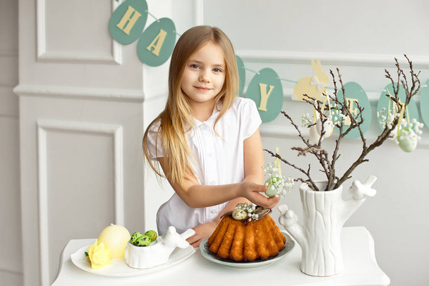 Beautiful blonde little girl in a white shirt is sitting at the holiday table near a vase with sprigs decorated with Easter eggs. - Fotoğraf, Görsel