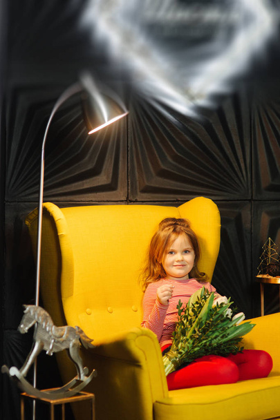 Маленькая девочка сидит на желтом кресле с букетом белых тюльпанов
 - Фото, изображение