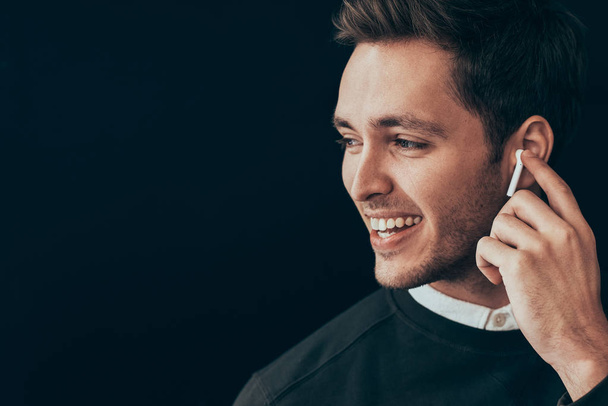 Closeup horizontal portrait of young handsome man smiling and have a call with a colleague isolated on black background holding wireless earphone with finger. Businessman using wireless headphone. - Foto, Imagen