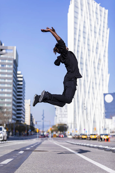 Side view of a young black man wearing casual clothes jumping in urban background. Lifestyle concept. Millennial african guy wearing sunglasses outdoor - Foto, afbeelding