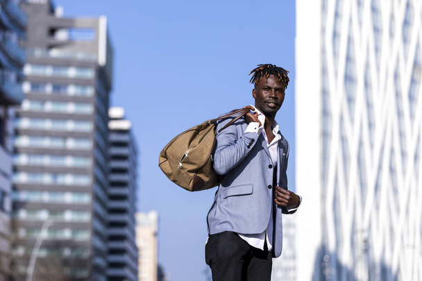 Front view black young man urban casual fashion in the street. Wearing black elegant jacket, jeans, holding and carrying a bag in a sunny day - 写真・画像