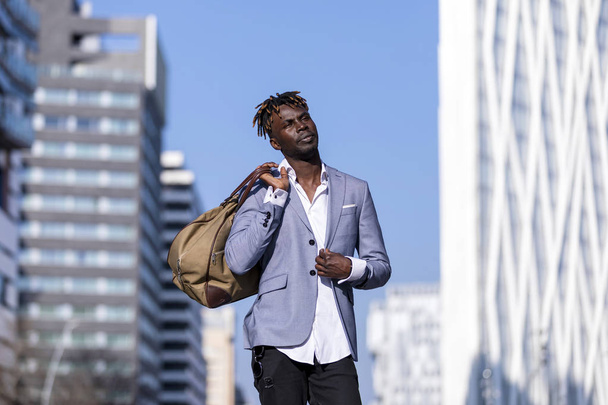 Frontansicht schwarzer junger Mann in urbaner Freizeitmode auf der Straße. trägt schwarze elegante Jacke, Jeans, hält und trägt eine Tasche an einem sonnigen Tag - Foto, Bild