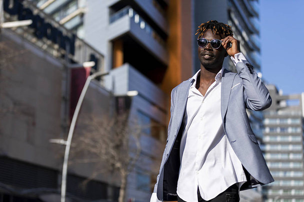 Low angle view of a black african young man wearing elegant clothes with sunglasses put in on eyeglasses while walking on the street in a sunny day - Foto, afbeelding