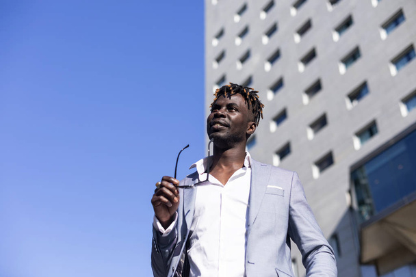 Low angle view of black african young man wearing elegant clothes with sunglasses put in on eyeglasses while walking on the street in a sunny day - 写真・画像