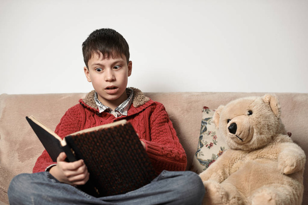 Мальчик читает книгу, сидит на диване с медвежьей игрушкой
 - Фото, изображение