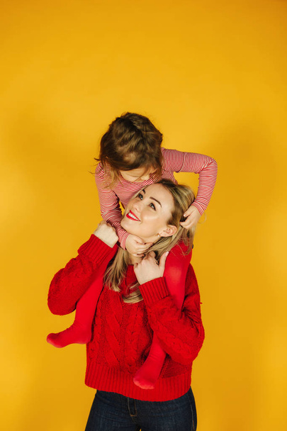 Happy little girl with her young mother on the yellow background. Daughter sitting on moms neck. Smile - Foto, Imagen
