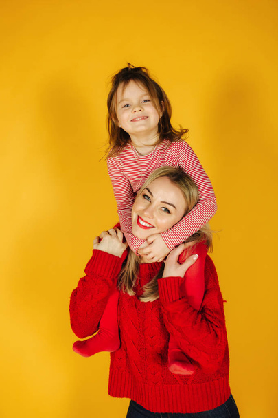 Gelukkig klein meisje met haar jonge moeder op de gele achtergrond. Dochter zit op moeders nek. Glimlach - Foto, afbeelding