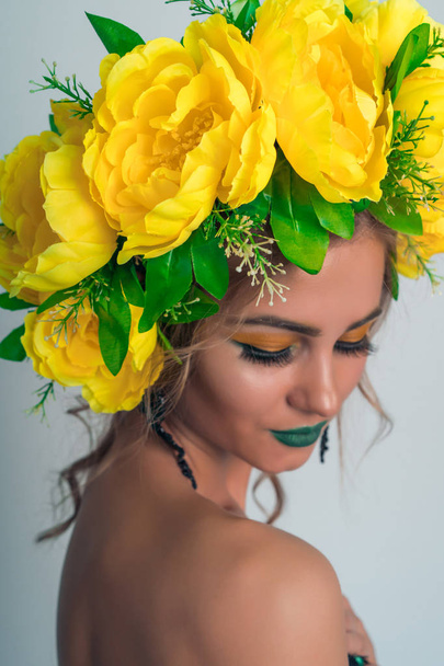 Retrato elegante de uma linda garota loira com uma coroa de flores amarelas grandes em sua cabeça e maquiagem brilhante em um fundo cinza
 - Foto, Imagem