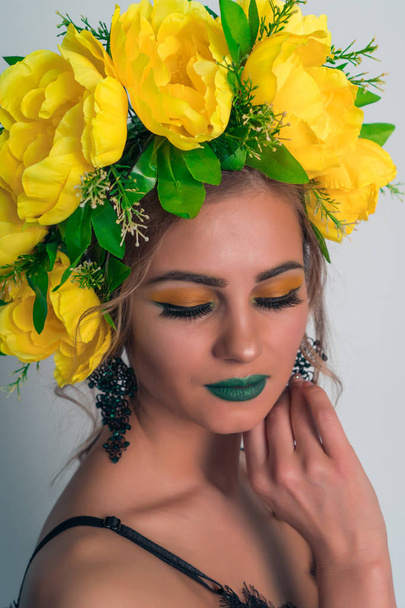 Fashionable portrait of a beautiful blonde girl with a wreath of large yellow flowers on her head and bright makeup on a gray background - Photo, image
