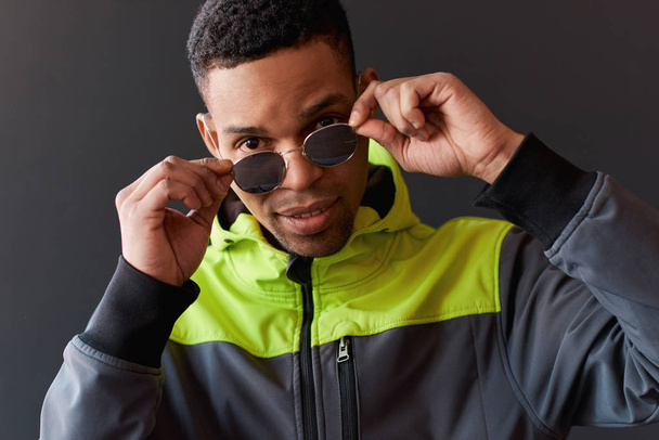Closeup portrait of stylish African American man looking through posing for mirror sunglasses, posing on black studio wall. People, emotion and lifestyle concept - Foto, imagen