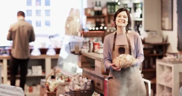パンの塊を保持しているエプロンを身に着けているデリカテッセン ショップの女性オーナーの笑顔のポートレート - 映像、動画